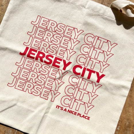 canvas tote bag that repeatedly has Jersey City written across it in red letters. Under Jersey City it reads It's a Nice Place.