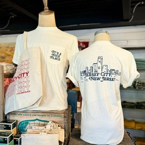 Two mannequins wearing the the same shirt. One pictures the front of the white tee shirt with a right breast pocket. On the pocket reads "It's A Nice Place" in black letters. The other mannequin shows the back of the shirt and it reads "Jersey City, New Jersey" with an outline of the city skyline.