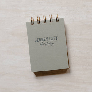 A gray rectangular mini jotter notepad that reads "Jersey City, NJ" on the cover